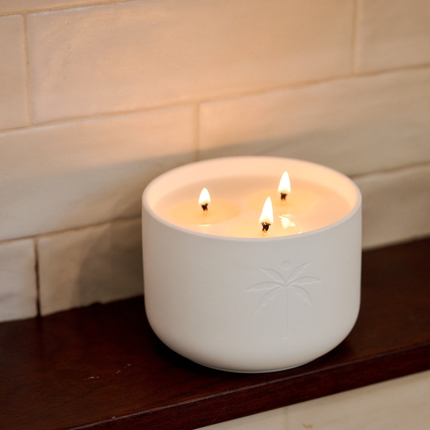 White ceramic candle holder with multiple lit candles on a wooden shelf against a tiled wall.